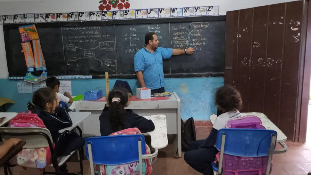 Un maestro de pie junto a un pizarrón con palabras escritas enseña a alumnos sentados en sus pupitres.