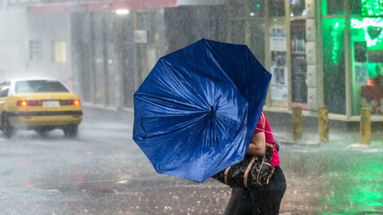 Persona con paraguas azul bajo una intensa lluvia en la calle mientras pasa un taxi amarillo.