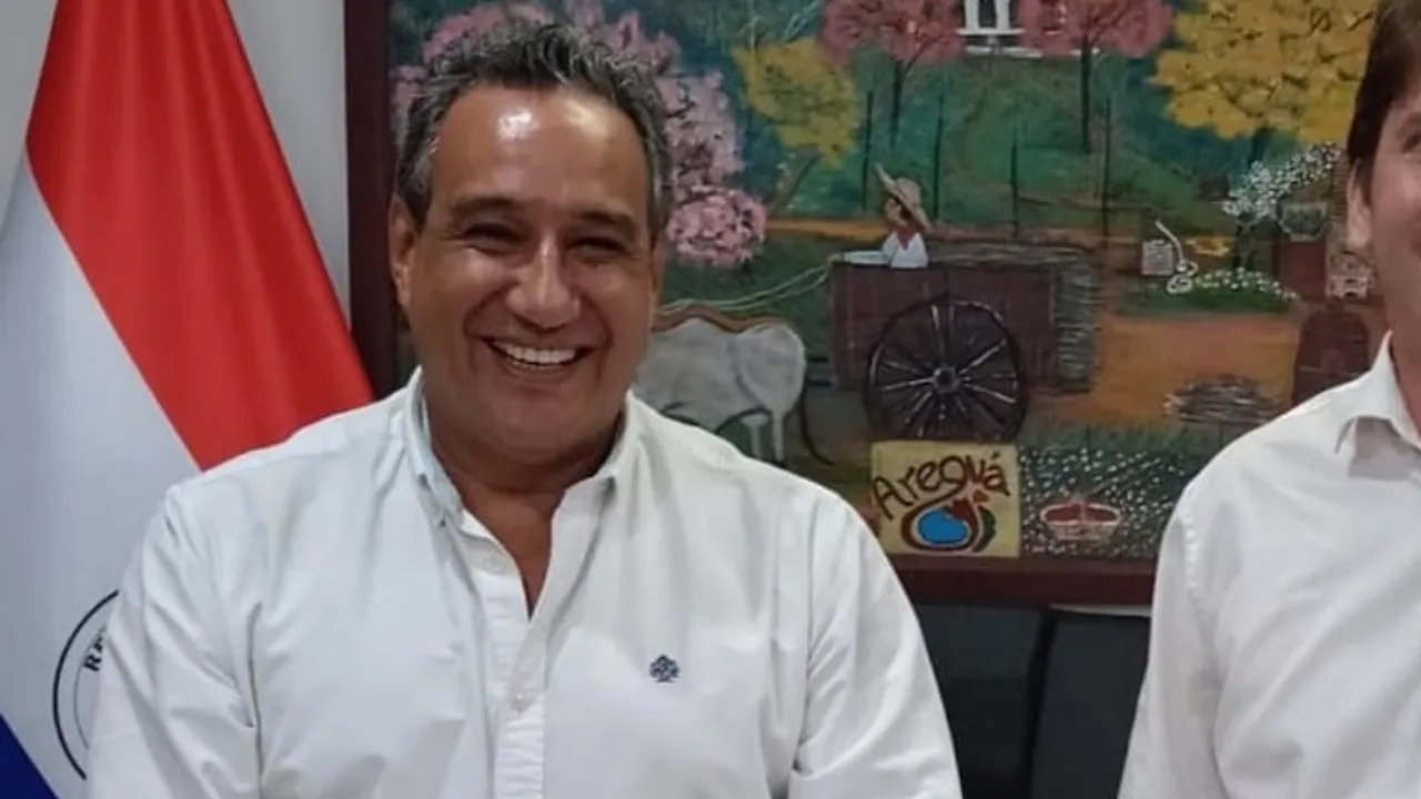 Hugo Javier González sonríe vistiendo una camisa blanca, con la bandera paraguaya a la izquierda.