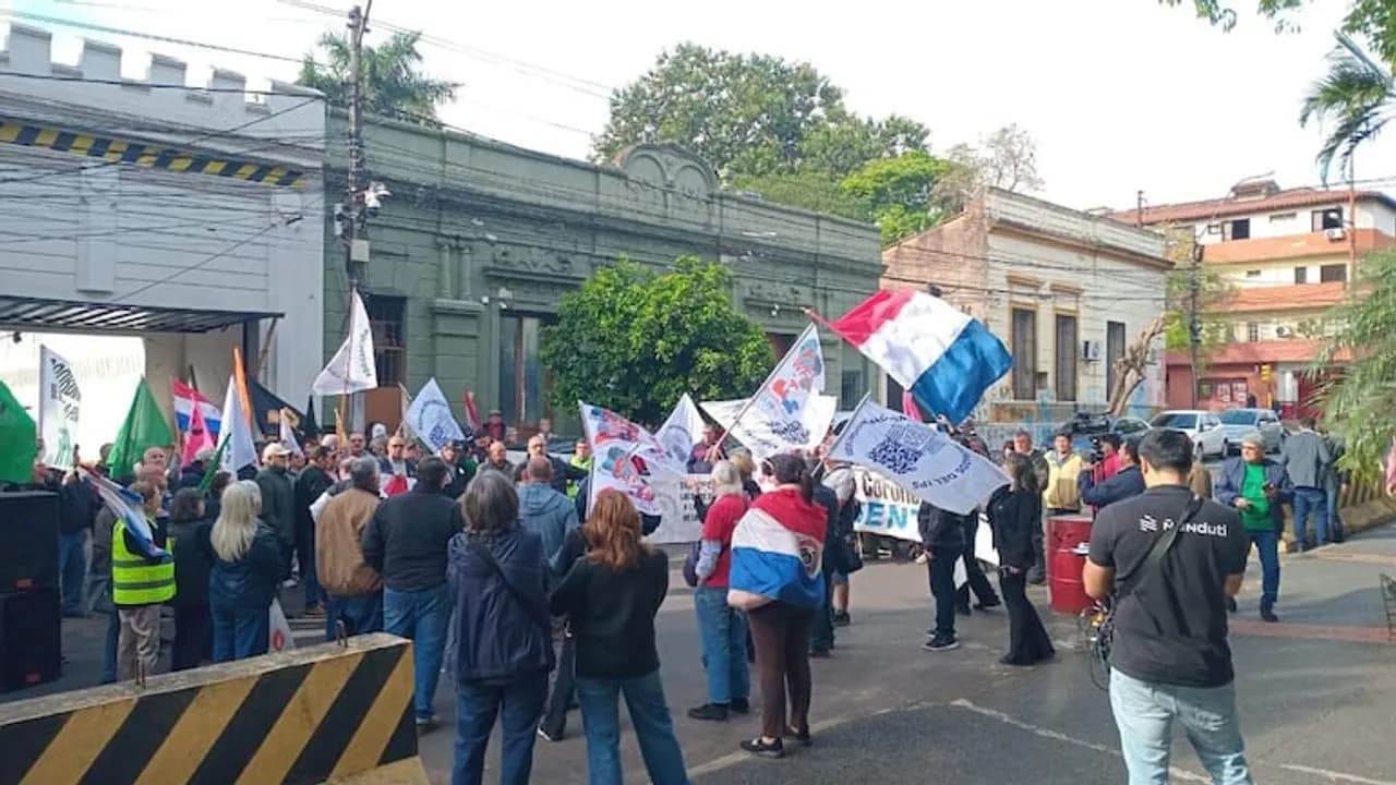 Grupo de personas manifestándose en la calle con banderas de Paraguay y carteles frente a edificios antiguos.