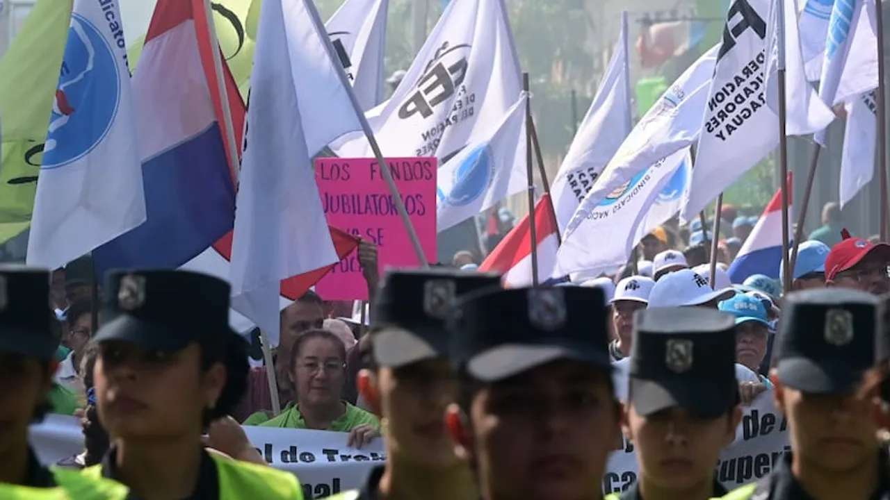 Manifestantes marchan con banderas paraguayas y de gremios docentes detrás de una fila de policías uniformados.