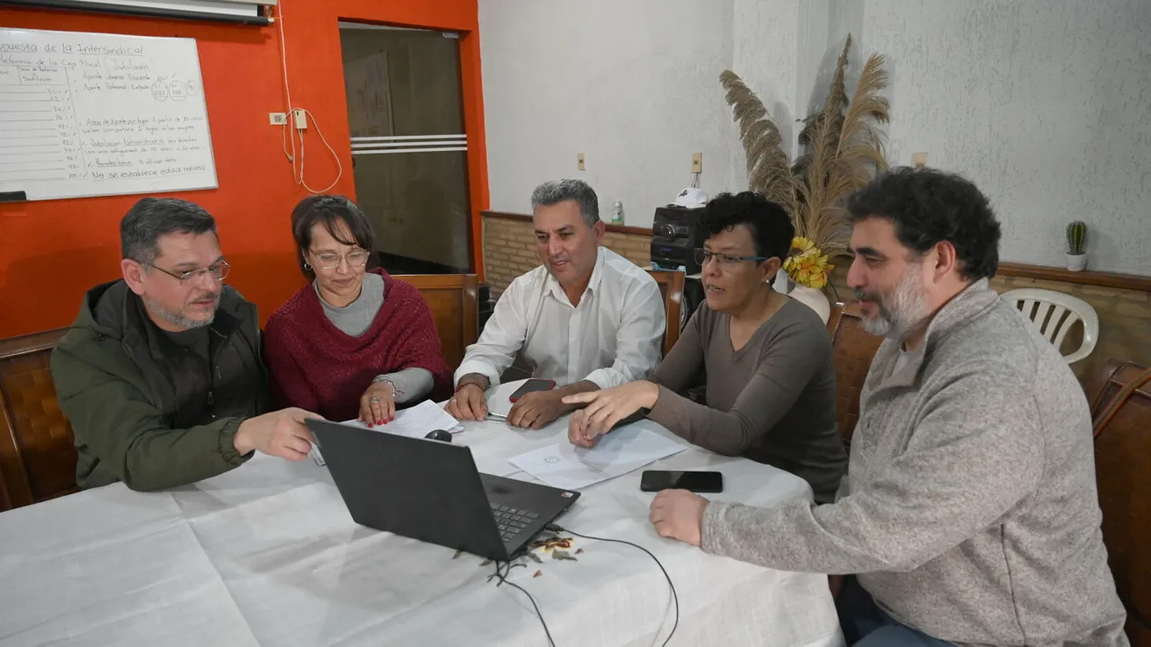 Cinco personas reunidas alrededor de una mesa analizan documentos frente a una computadora portátil.