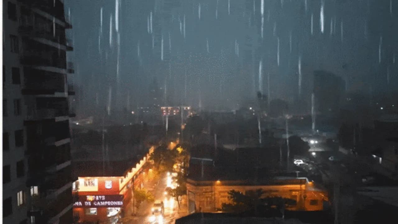 Vista nocturna de una calle de la ciudad bajo una intensa lluvia con luces de edificios y vehículos.