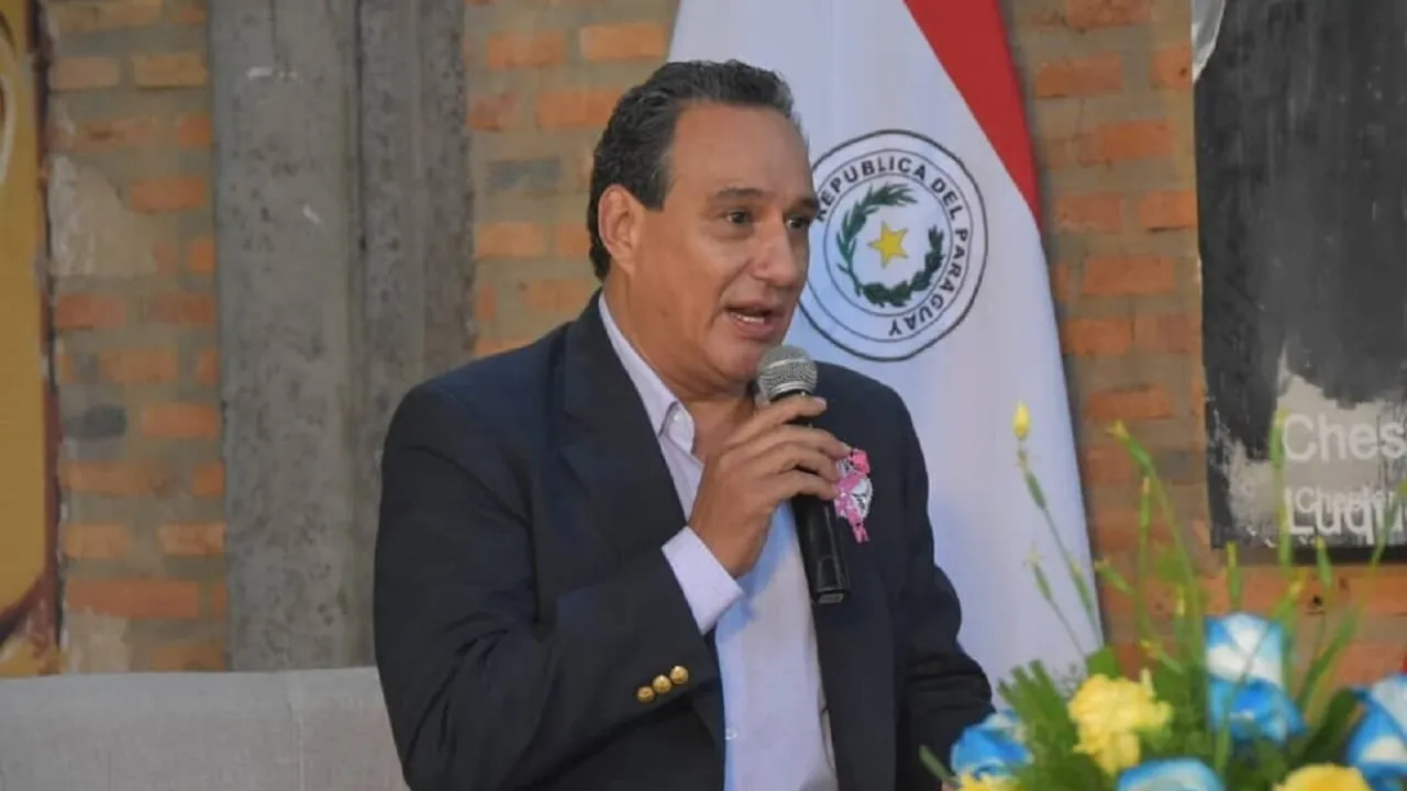Hugo Javier González habla frente a un micrófono, vistiendo un saco oscuro, con la bandera de Paraguay de fondo.