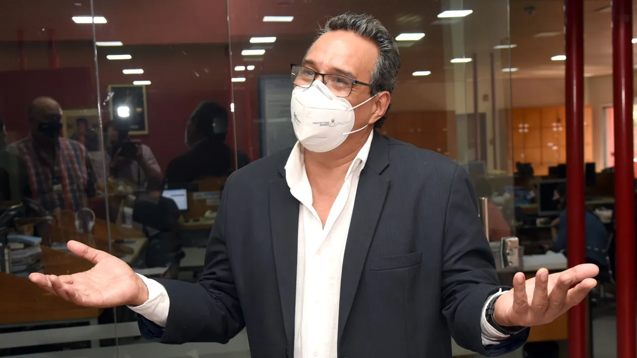 Hugo Javier González con tapabocas blanco, anteojos y saco oscuro, gesticulando con las manos abiertas.