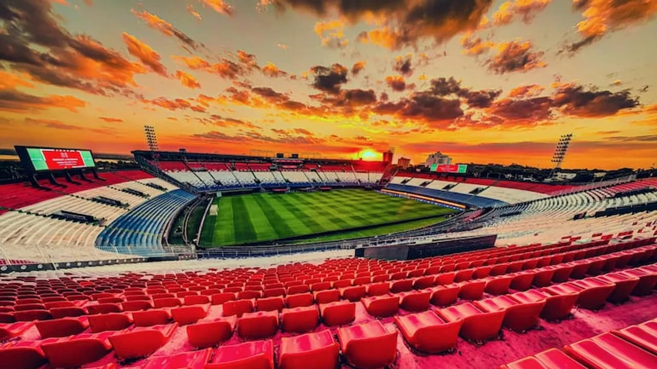 Vista panorámica de las graderías vacías del estadio Defensores del Chaco bajo un cielo atardecido con nubes naranjas.