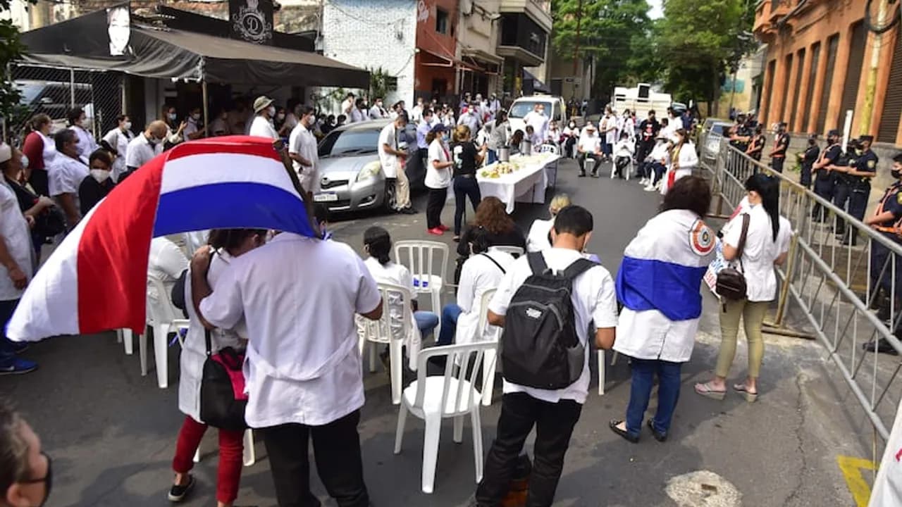 Personal de blanco con banderas paraguayas se manifiesta en una calle frente a una barrera policial.