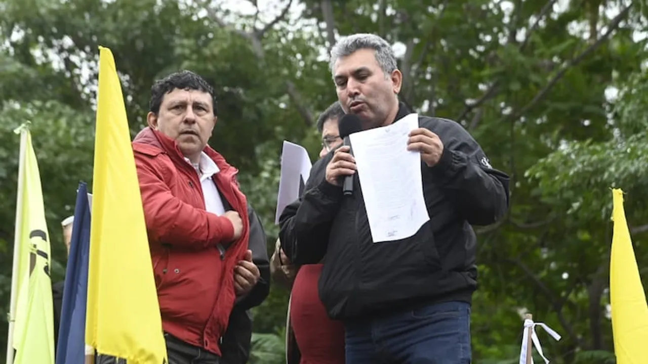 Silvio Piris habla por micrófono sosteniendo un documento junto a manifestantes con banderas amarillas.