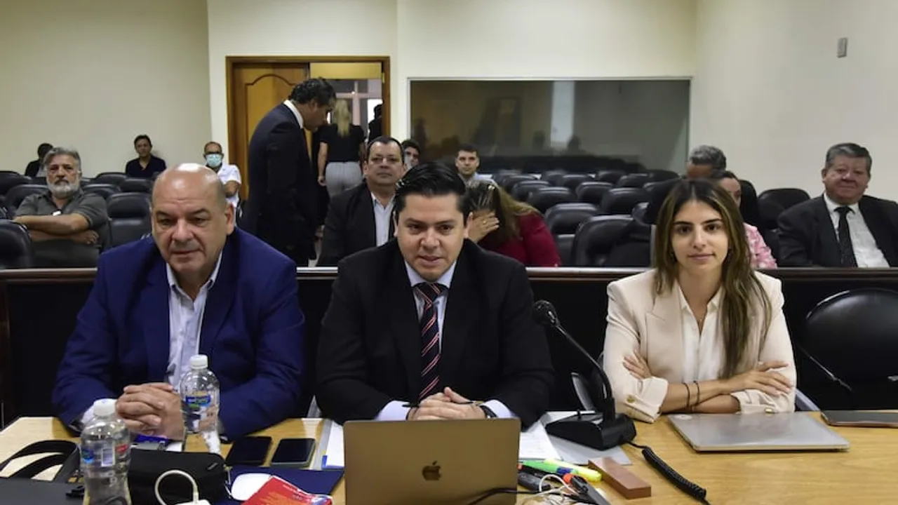 Tres personas sentadas frente a un escritorio con una computadora en una sala de audiencias.