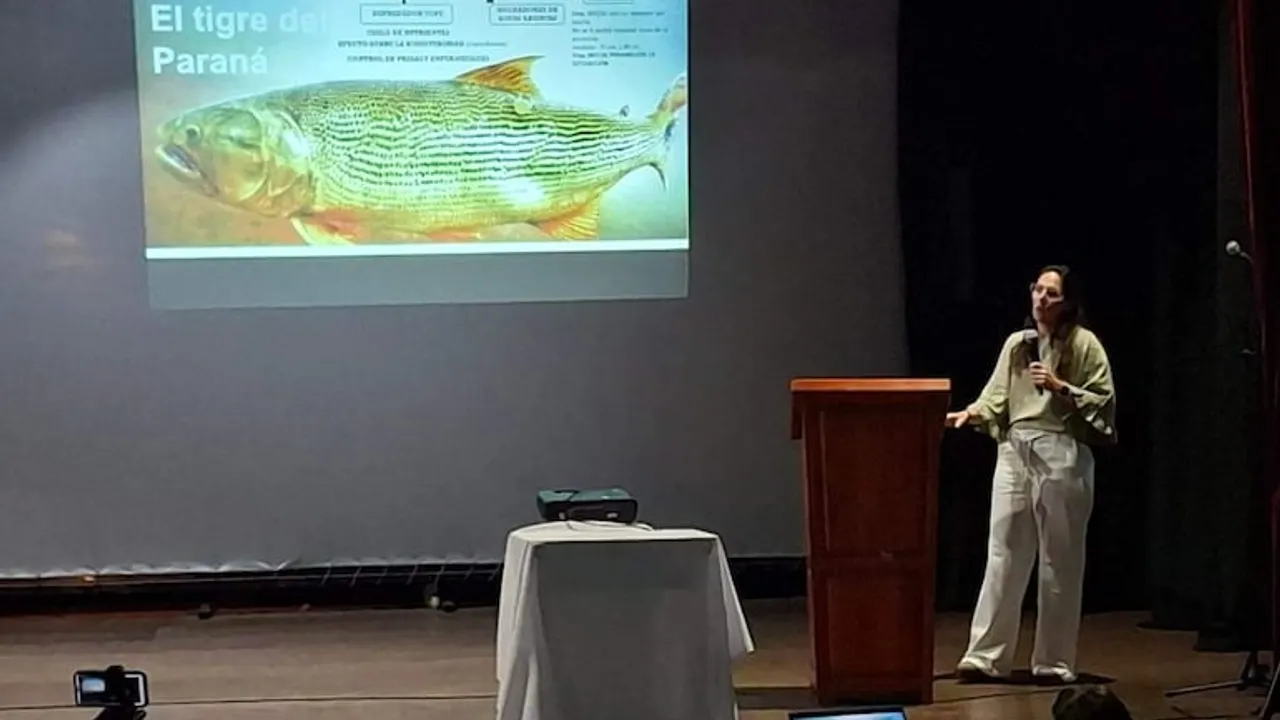 Una mujer expone en un escenario junto a una pantalla que muestra la imagen de un pez dorado.