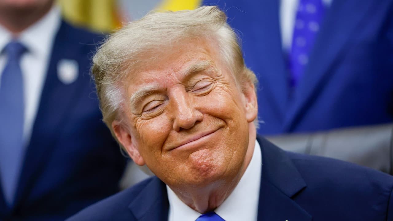 Donald Trump sonríe con los ojos cerrados vistiendo un traje azul oscuro y corbata, con personas de fondo.