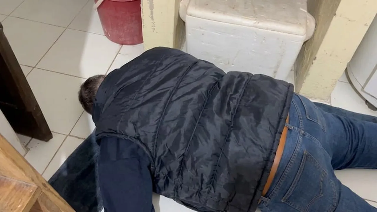 Hombre acostado boca abajo en el suelo de baldosas, vistiendo chaleco negro y jeans, junto a una conservadora.