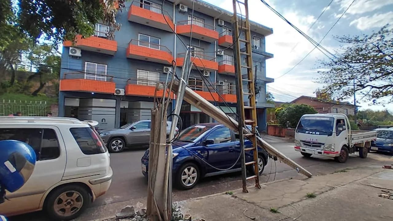 Poste de concreto inclinado sobre una calle, sostenido por una escalera de madera y cables.