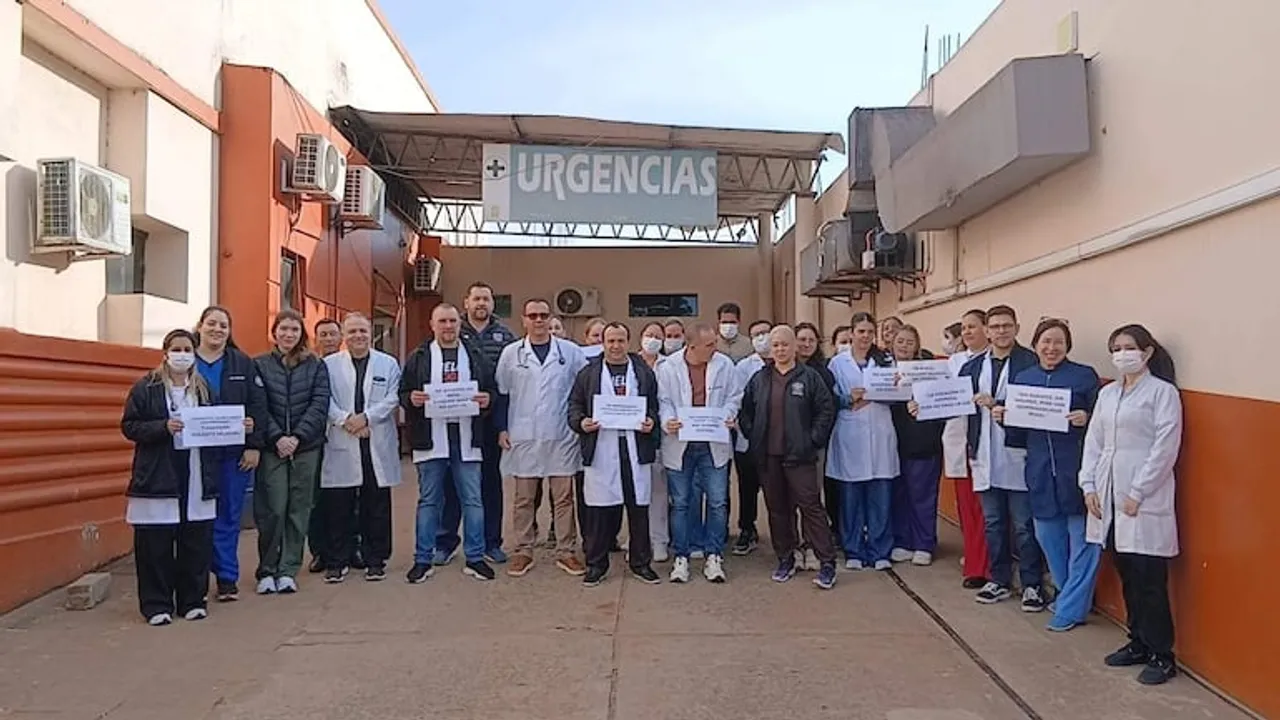Personal médico con batas blancas y uniformes posa con carteles de protesta frente al acceso de urgencias del hospital.
