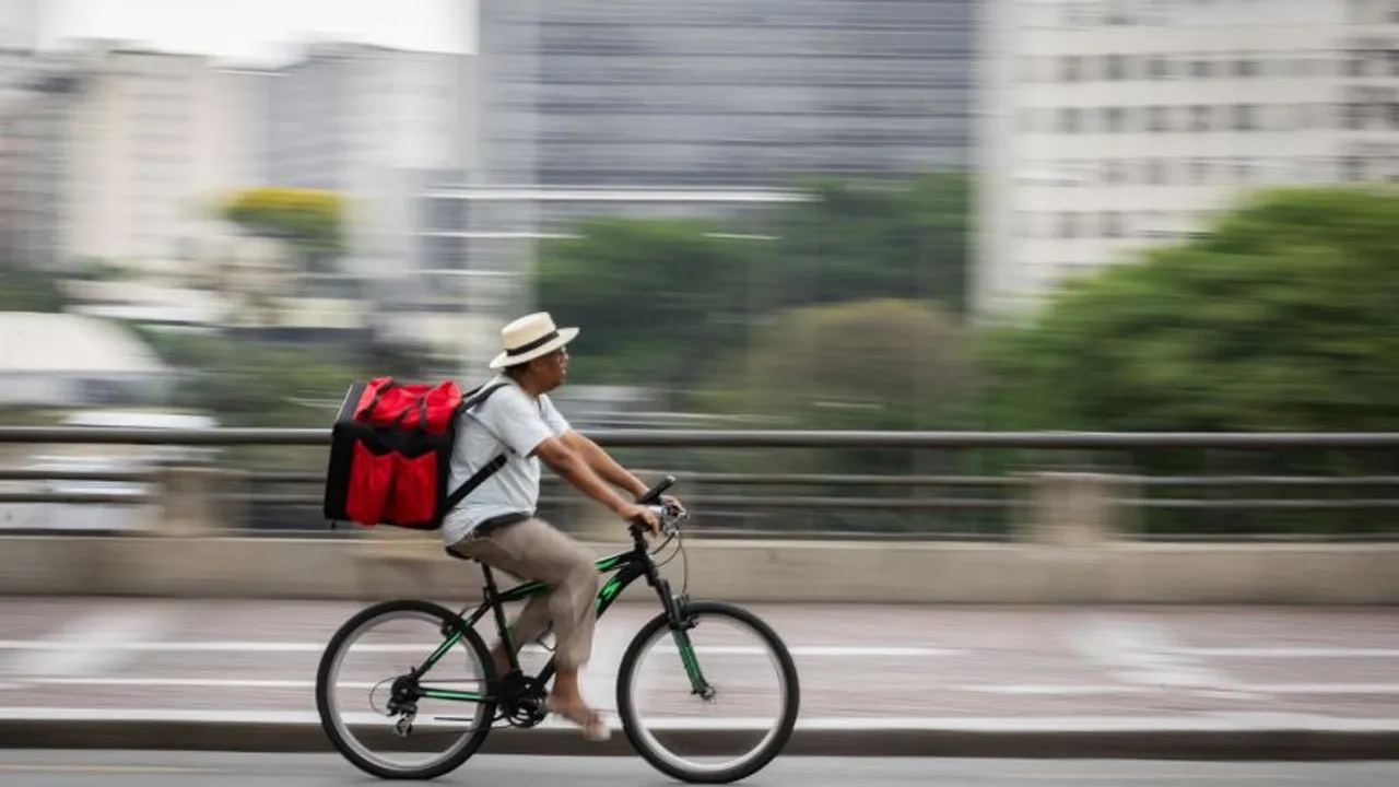 Repartidor con sombrero y una gran mochila roja pedalea en bicicleta por una calle urbana con efecto de movimiento.