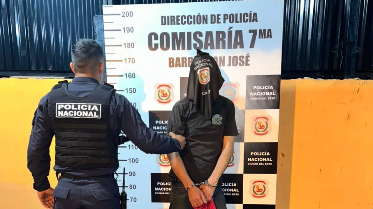 Un policía de espaldas sujeta a un detenido esposado con la cabeza cubierta frente a un cartel de la Comisaría Séptima.
