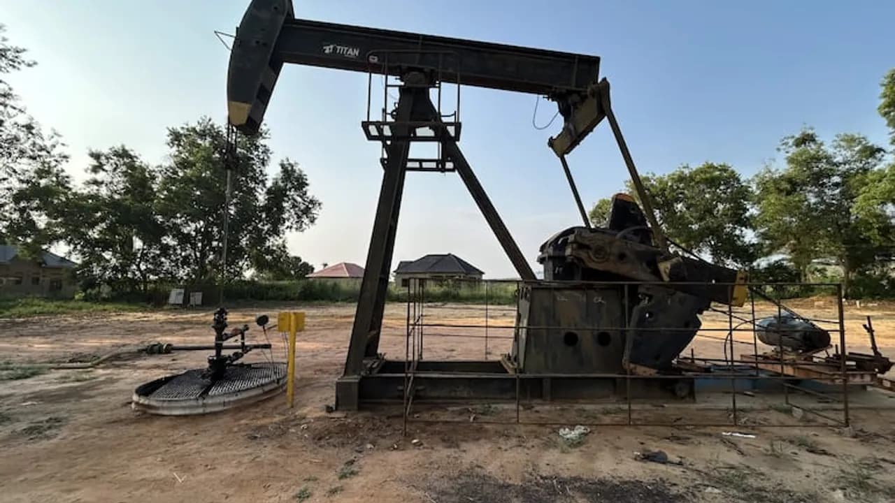 Bomba de extracción de petróleo de color negro en un terreno de tierra con árboles y casas al fondo.