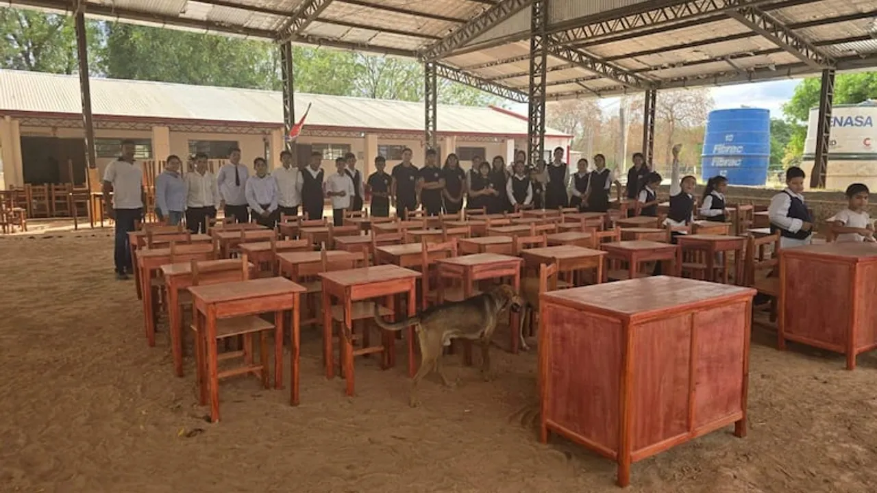 Estudiantes y adultos bajo un tinglado con piso de tierra junto a pupitres y escritorios de madera recién donados.