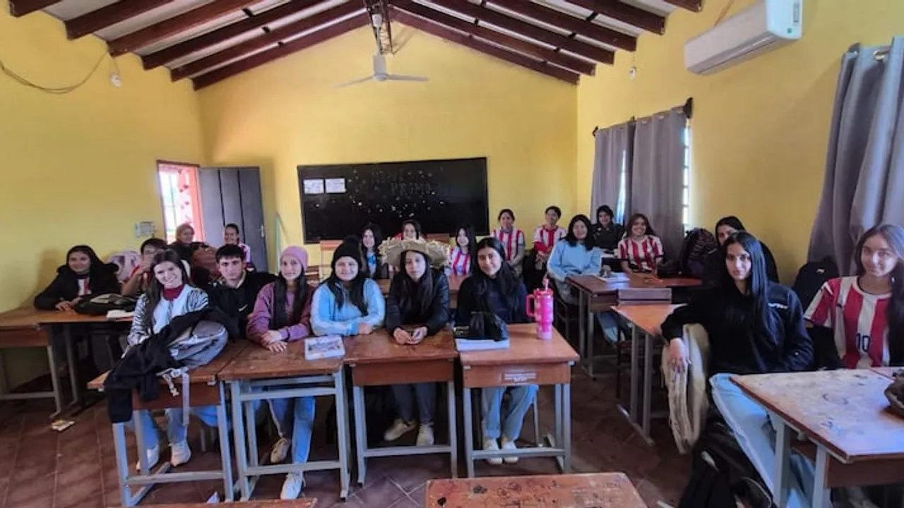 Estudiantes sentados en pupitres de madera dentro de un aula con paredes amarillas y techo de vigas.