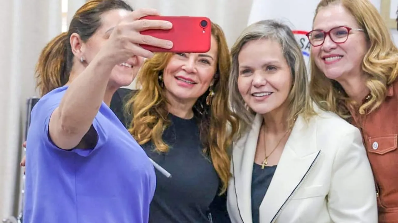 Cuatro mujeres sonríen juntas mientras una de ellas toma una selfie con un teléfono celular rojo.