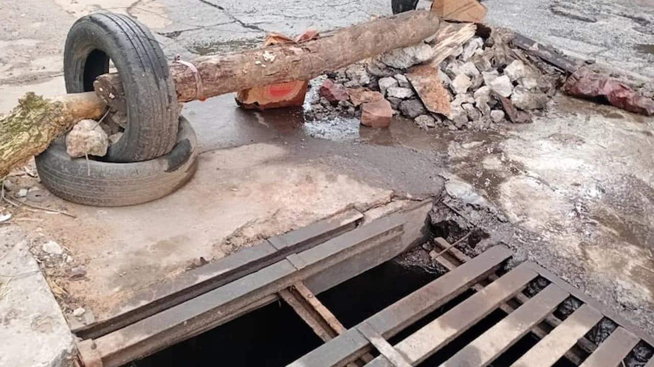 Tronco de madera colocado dentro de neumáticos y piedras para señalizar un pozo peligroso junto a una rejilla de metal.