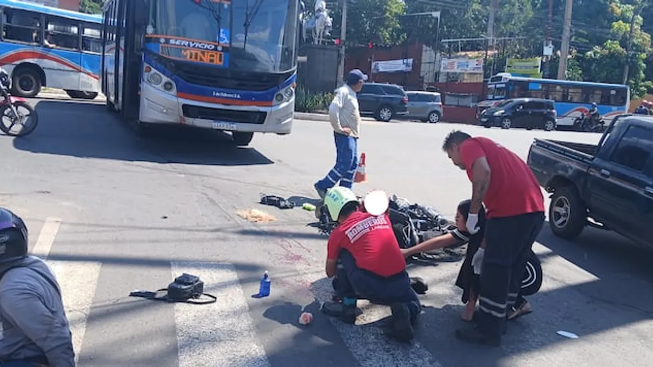 Bomberos asisten a una persona tendida en el asfalto junto a una moto, frente a un bus de la empresa 3 de Febrero.
