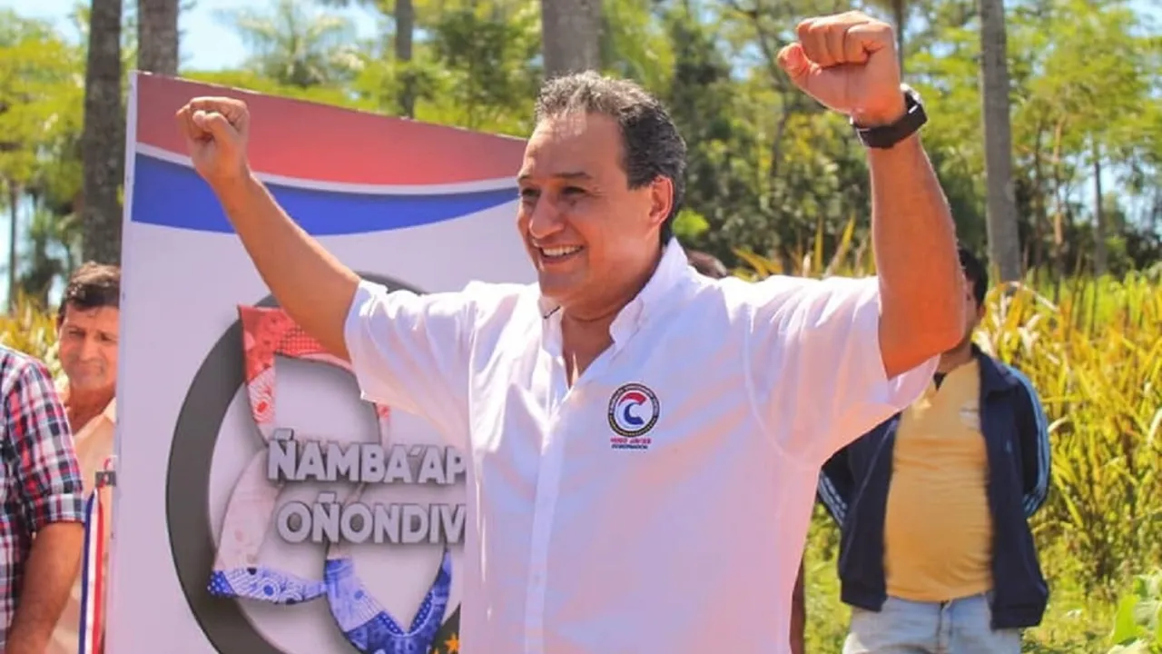 Un hombre sonriente con camisa blanca levanta ambos puños en señal de victoria frente a un cartel.