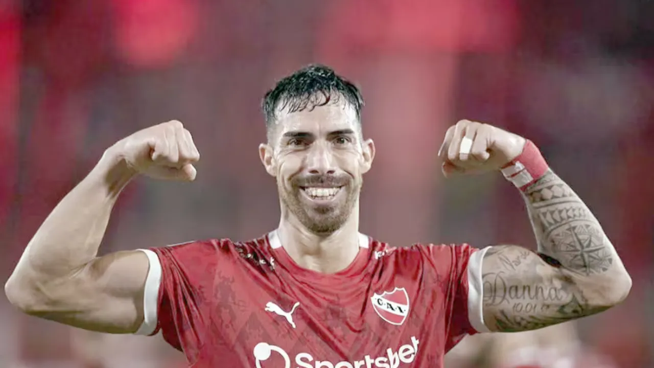 Gabriel Ávalos sonríe y levanta ambos brazos flexionados, vistiendo una camiseta roja deportiva con tatuajes en el antebrazo.
