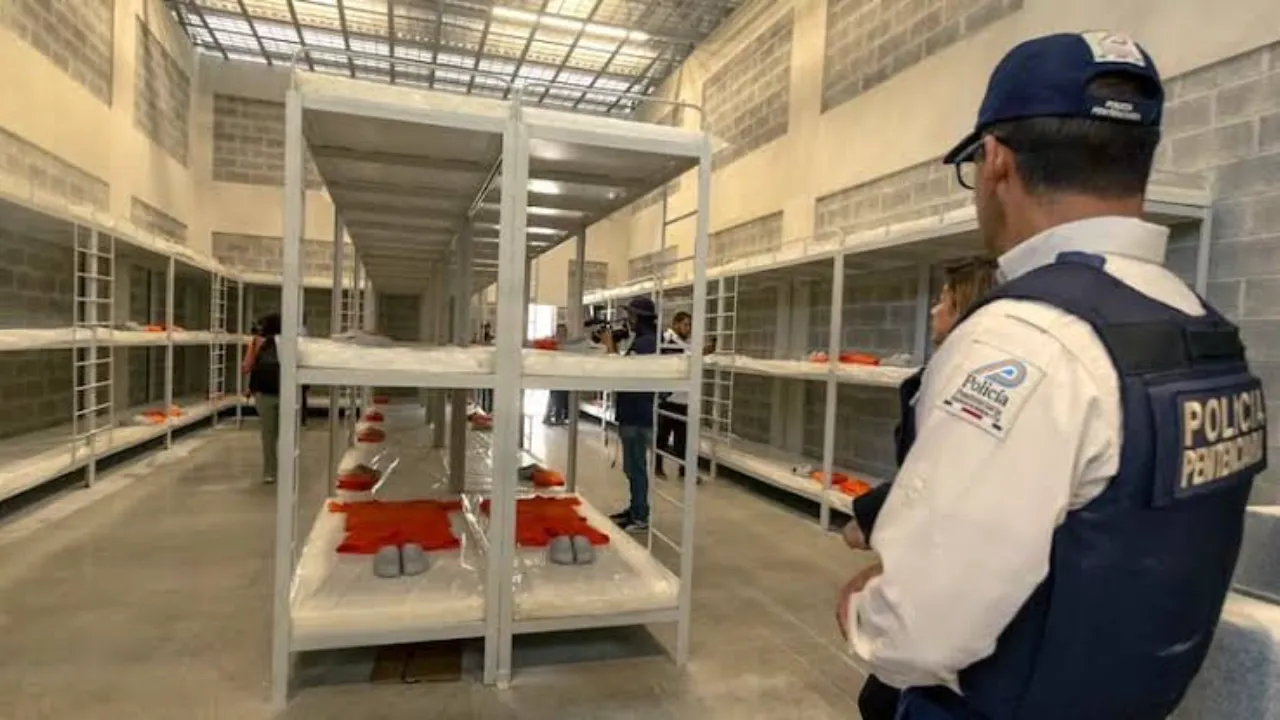 Oficial de seguridad custodia pasillo con literas metálicas y uniformes anaranjados en el penal de máxima seguridad.