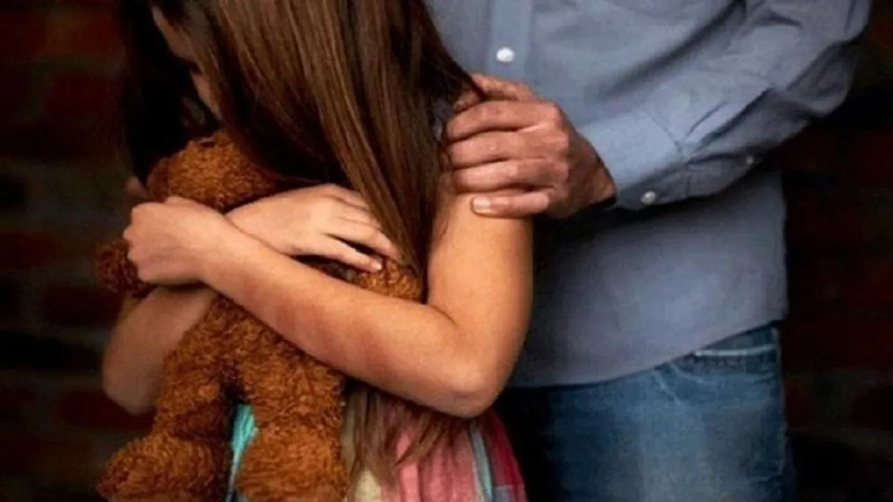 Niña abrazando un oso de peluche mientras una persona adulta apoya una mano sobre su hombro.