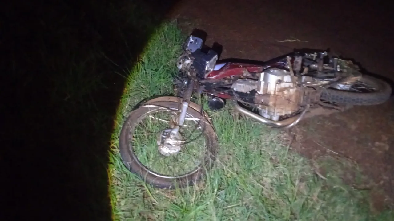 Motocicleta roja caída y destruida al costado de la ruta en la vegetación durante una noche oscura.