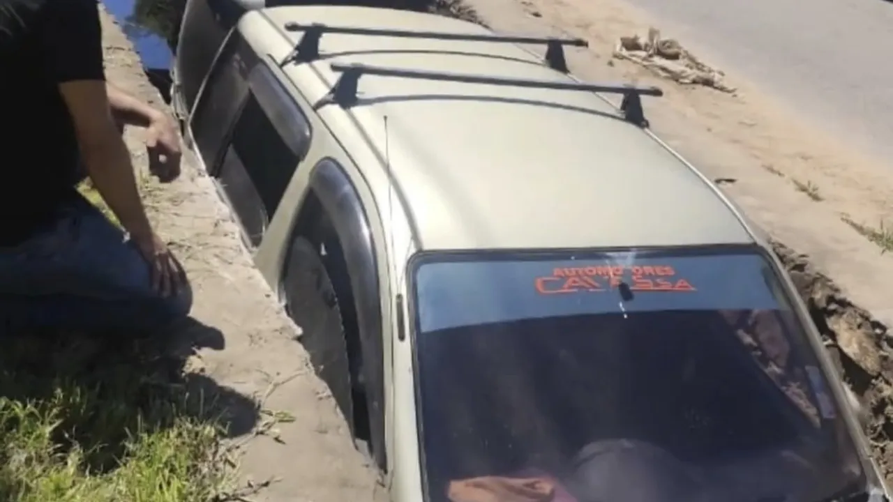 Vehículo volcado de costado dentro de una profunda canaleta de desagüe al costado de una calle asfaltada.