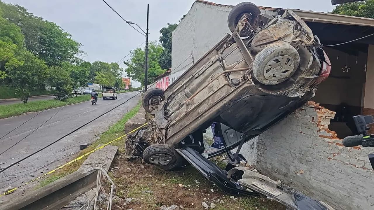 Automóvil volcado con las ruedas hacia arriba incrustado sobre la muralla dañada de una vivienda urbana.