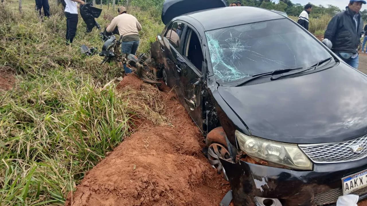 Automóvil negro con parabrisas roto caído en una zanja junto a una motocicleta destrozada y varias personas.