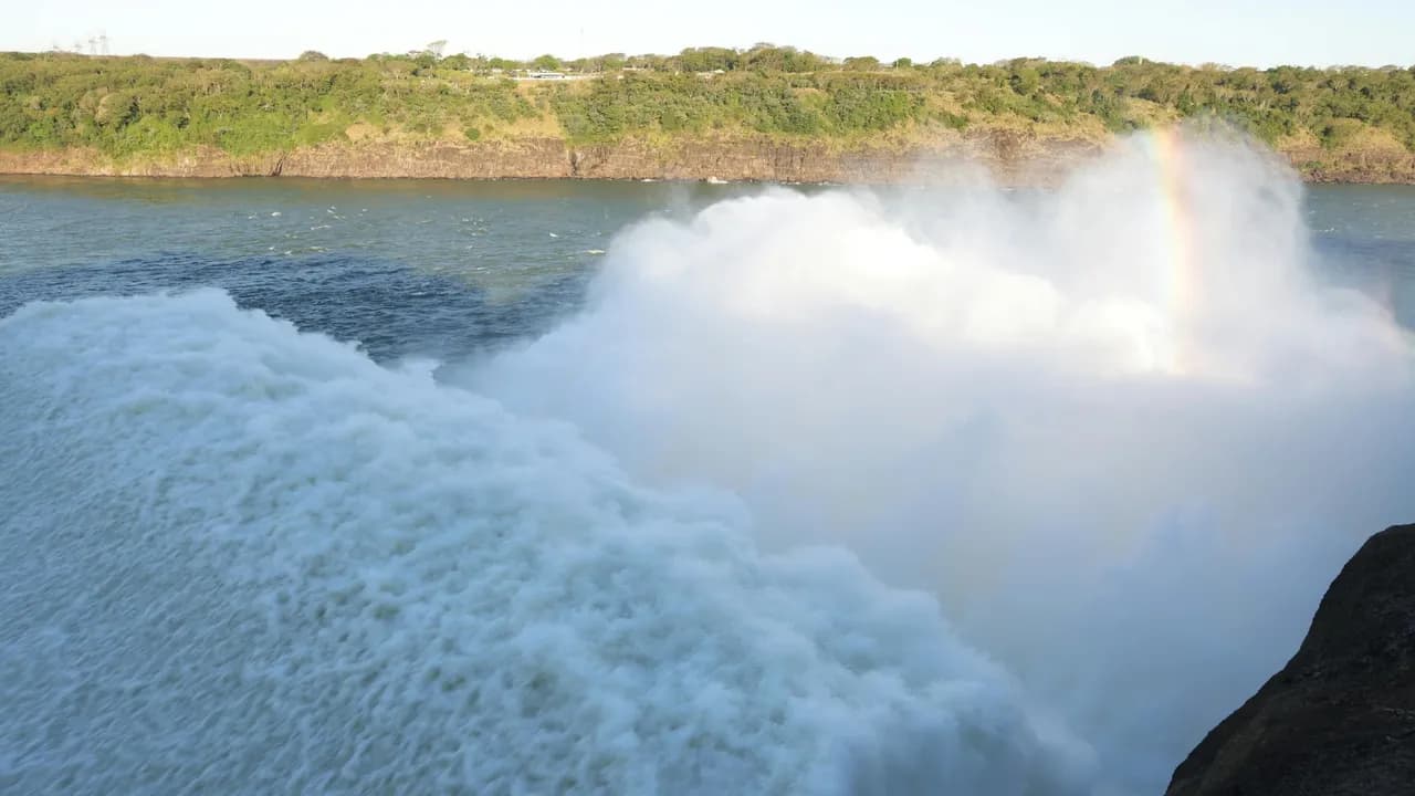 Agua saliendo con fuerza por las compuertas del vertedero de una represa hidroeléctrica, formando un arcoíris tenue.