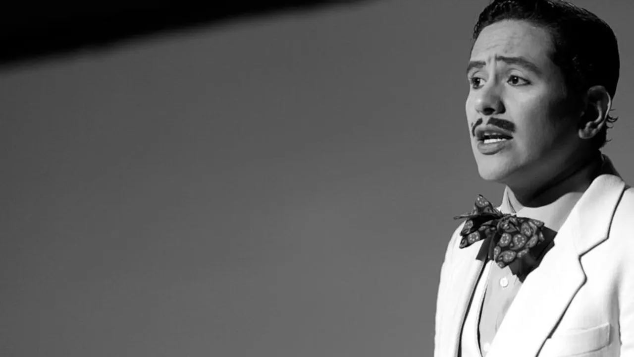 Hombre vestido con traje claro y moño actuando sobre un escenario teatral en una toma en blanco y negro.