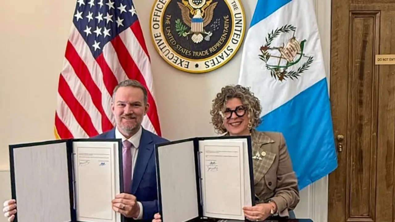 Dos autoridades presentan carpetas con acuerdos firmados frente a banderas oficiales de Estados Unidos y Guatemala.