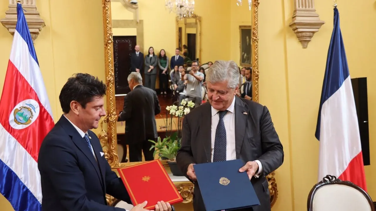 Dos representantes diplomáticos sostienen carpetas oficiales ante las banderas de Costa Rica y Francia