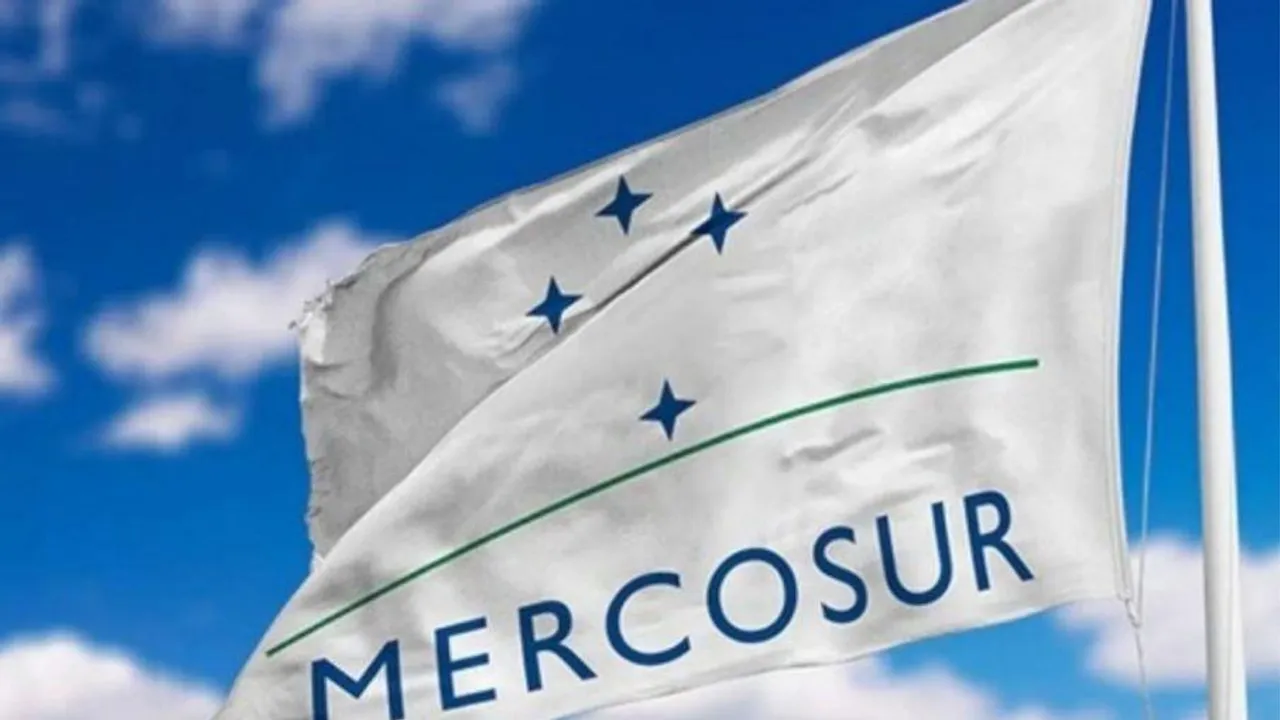 Bandera blanca oficial del bloque Mercosur flameando contra un cielo azul con nubes dispersas.
