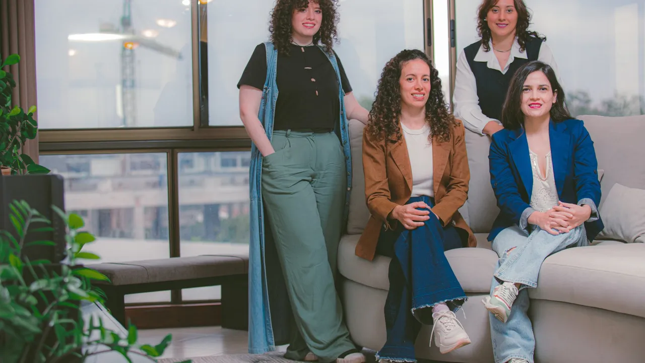 Cuatro mujeres profesionales del equipo de comunicación posan sonrientes en un sofá dentro de una oficina moderna.