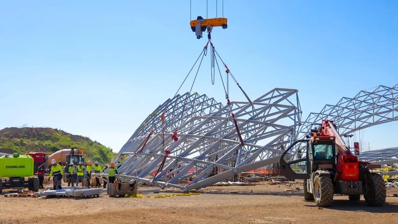 Grúa industrial levanta una estructura metálica curva en un predio en obras con operarios y maquinaria pesada.