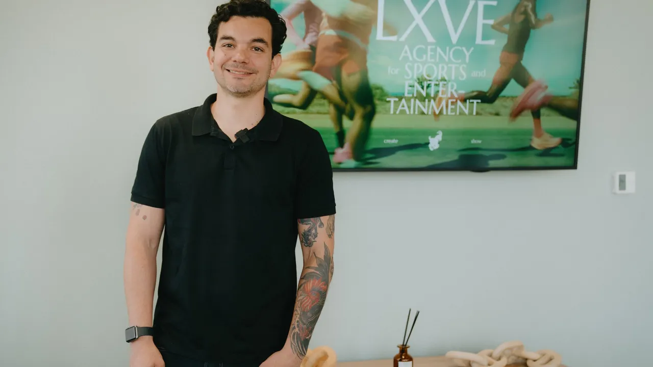 Jorge Tercarioli sonríe de pie con camisa polo negra frente a una pantalla con el logotipo de LXVE.