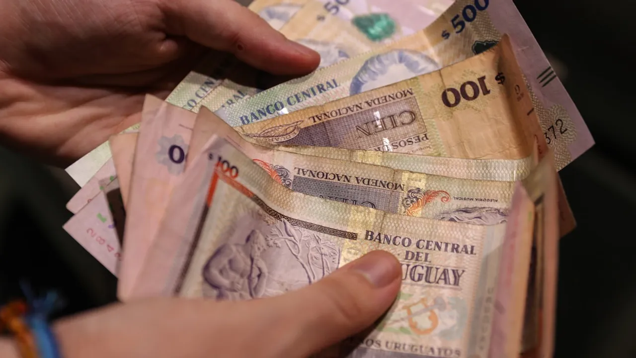 Manos sosteniendo varios billetes de cien y quinientos pesos uruguayos en efectivo sobre fondo oscuro.