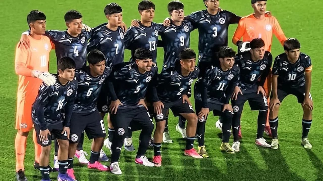 Plantel de futbolistas juveniles posando con indumentaria azul y arqueros de naranja en una cancha.