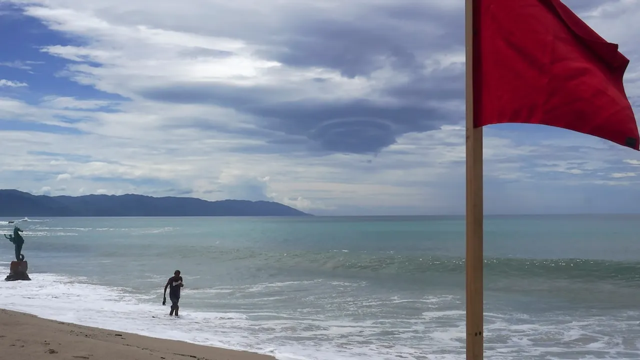 Bandera roja en la arena de una playa frente al mar con olas suaves y un cielo cubierto de densas nubes.