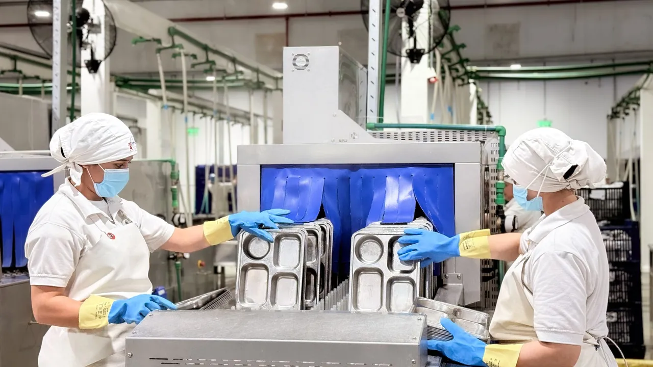 Dos trabajadoras con cofia y guantes manipulan bandejas metálicas para comida escolar en una línea industrial.