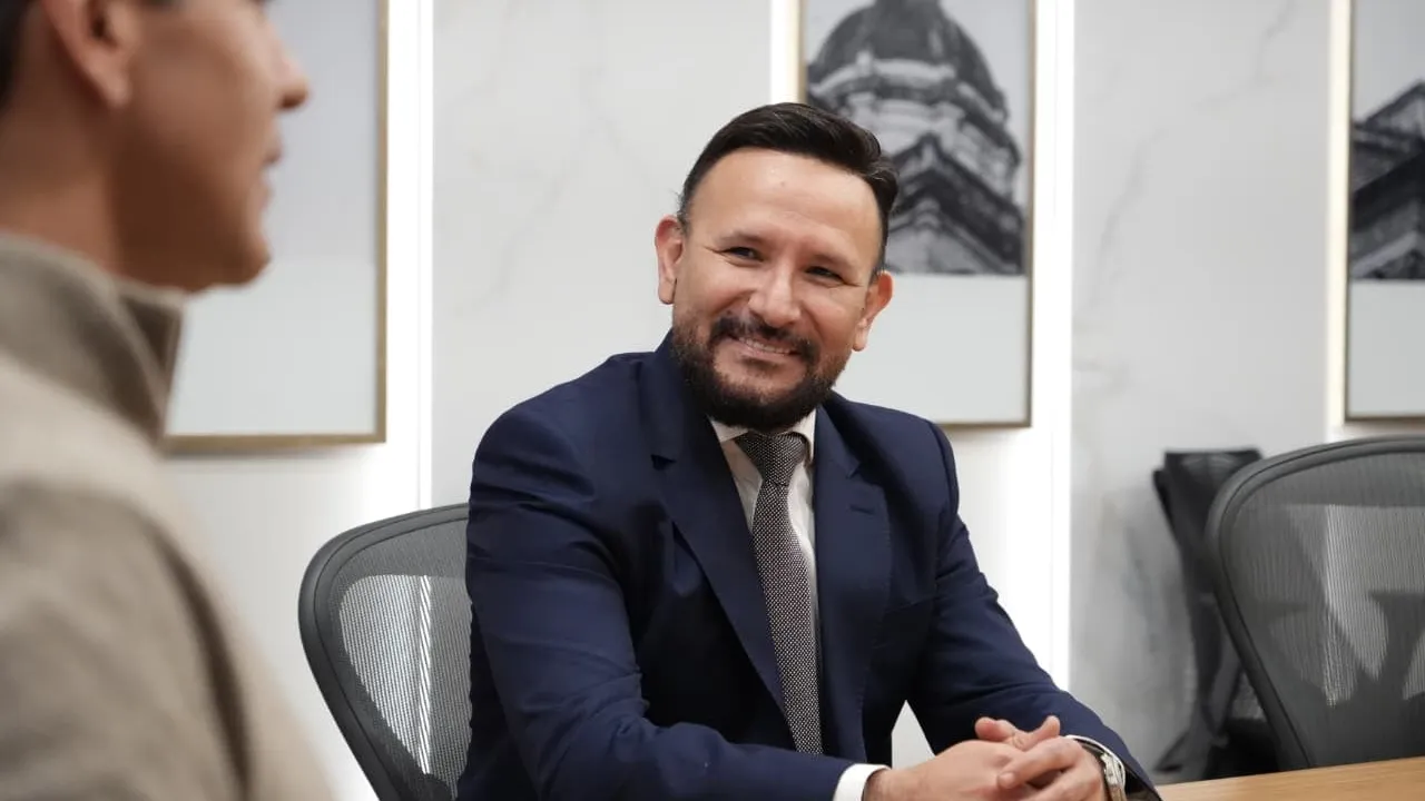 Hombre de traje azul marino y corbata sonríe sentado a una mesa de reuniones junto a un interlocutor.