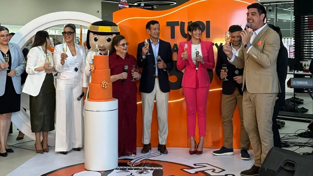 Directivos y mascota de la empresa brindan con copas de champaña junto a un pastel naranja por el aniversario.