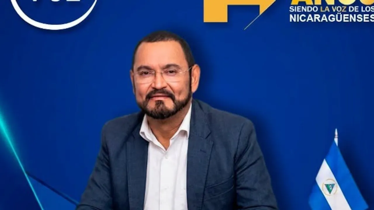 Hombre con barba recortada, gafas y saco azul oscuro sentado frente a un fondo azul con una pequeña bandera.