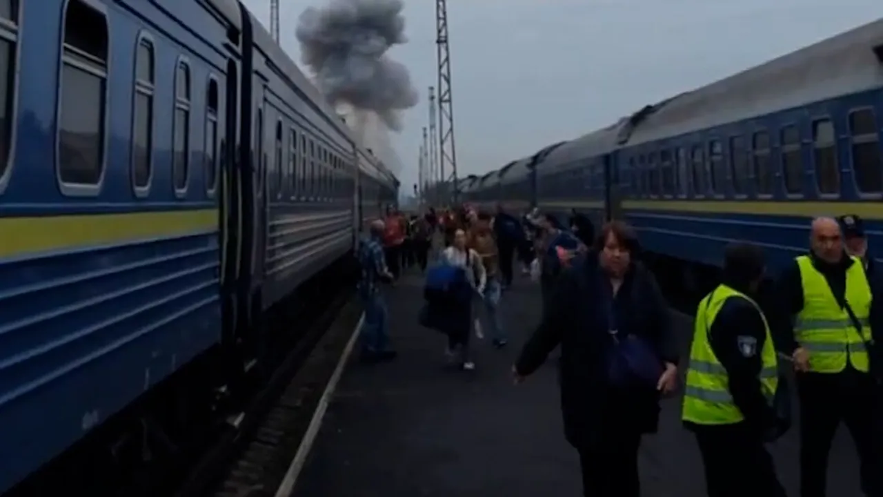 Pasajeros caminan por el andén entre dos trenes azules mientras una columna de humo se eleva a lo lejos.